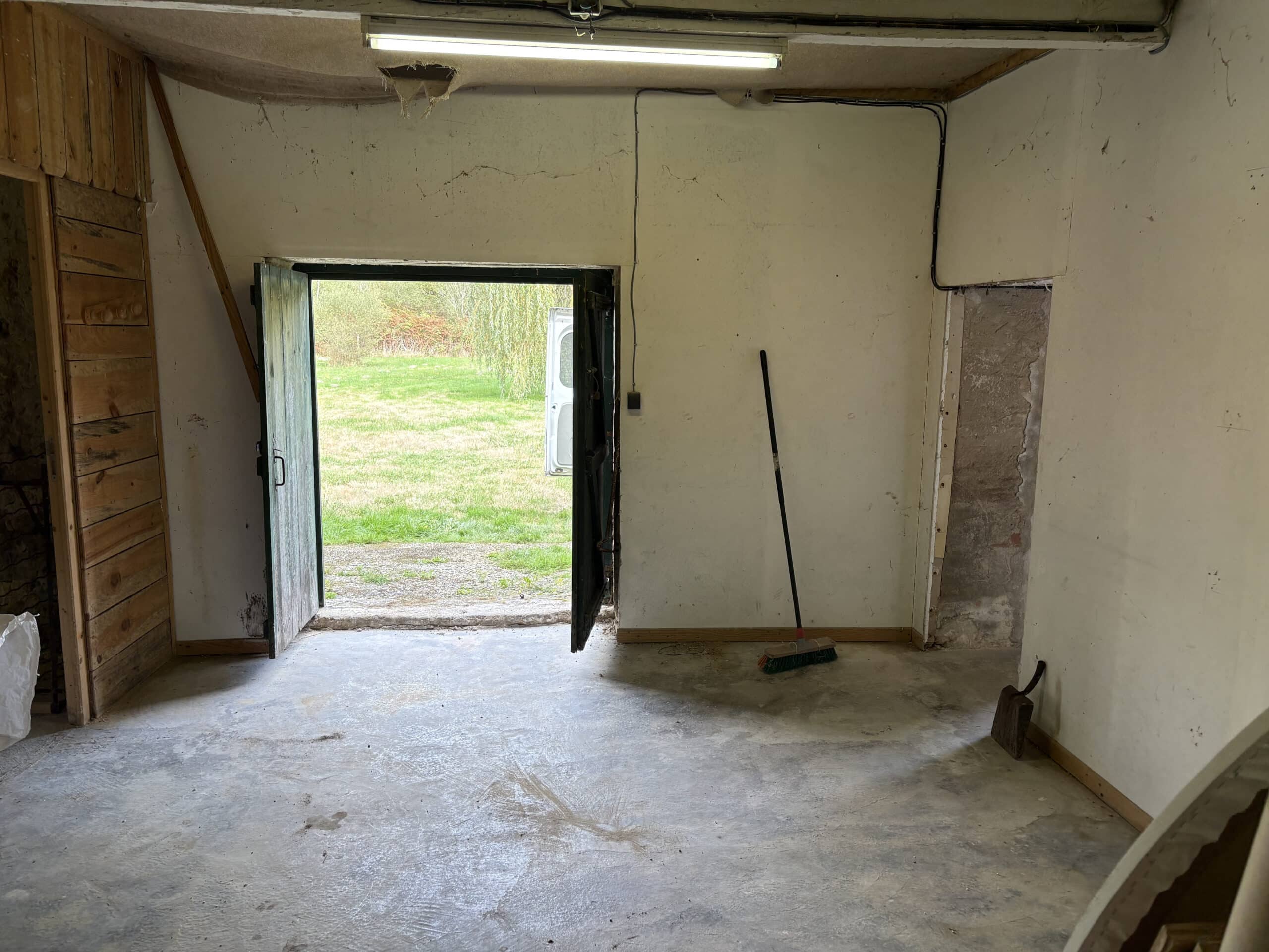 Intérieur d'un garage ou atelier vide, murs clairs abîmés, sol en béton, ouvrant sur une pelouse verte avec un véhicule blanc.