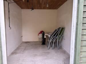 Intérieur d'un abri ou d'un petit espace de rangement avec sol en béton, plafond en bois et murs clairs. Chaises pliantes vertes et noires s'appuient contre le mur.