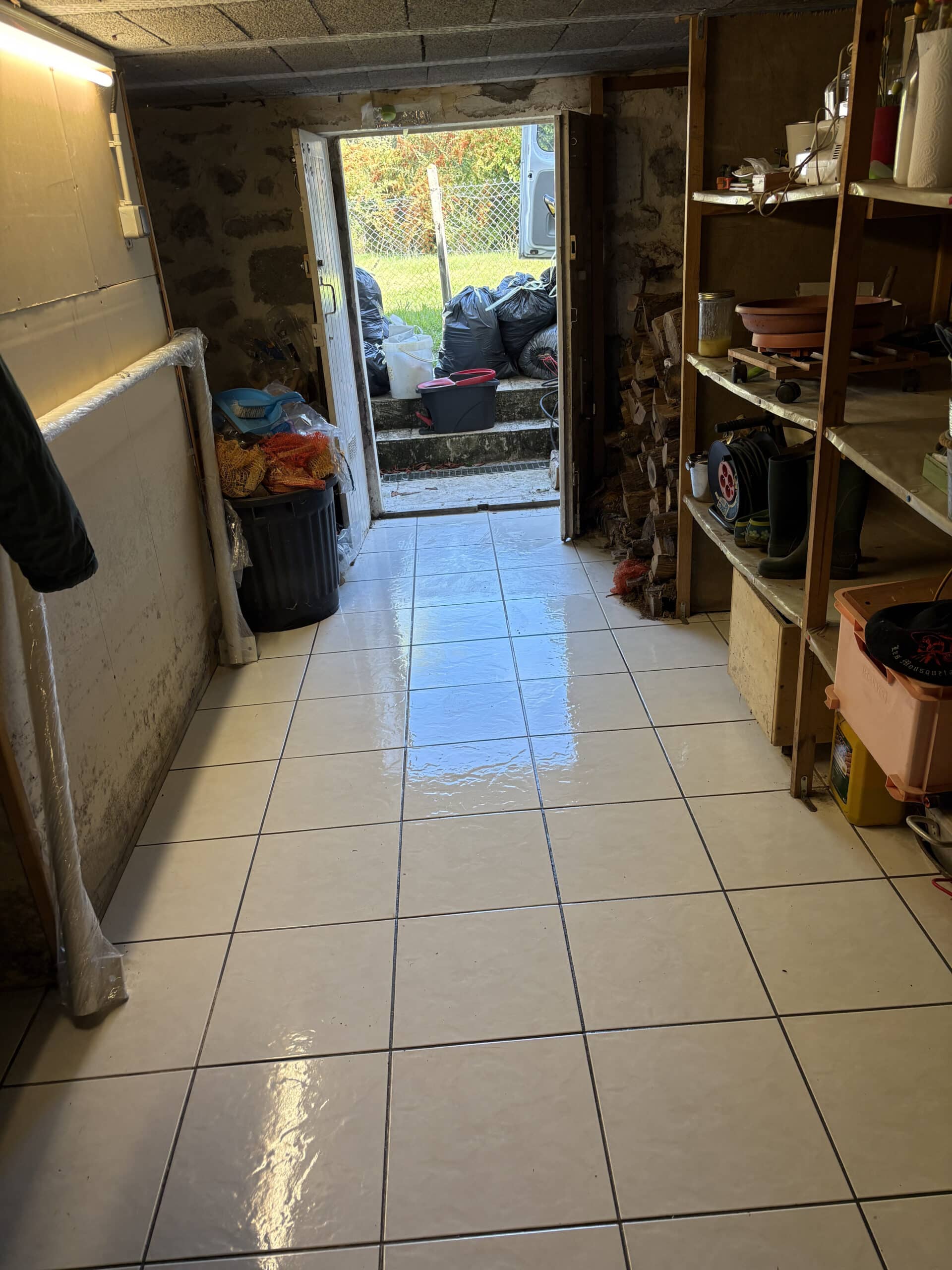 Entrée d'une pièce carrelée avec sol mouillé, menant à l'extérieur où s'entassent des sacs poubelles noirs près d'une camionnette.
