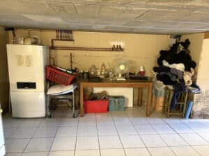 Chauffe-eau Buderus blanc dans une cave avec tuyauterie en cuivre, table, ventilateur UNITED, panier rouge et vêtements en tas.