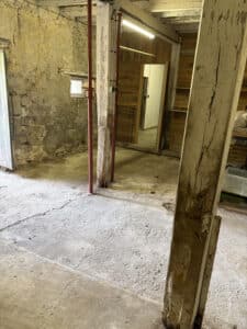 Intérieur rustique sombre d'une cave ou d'un grenier avec murs en pierre brute, sol en béton fissuré et poteaux de soutien temporaires rouges.