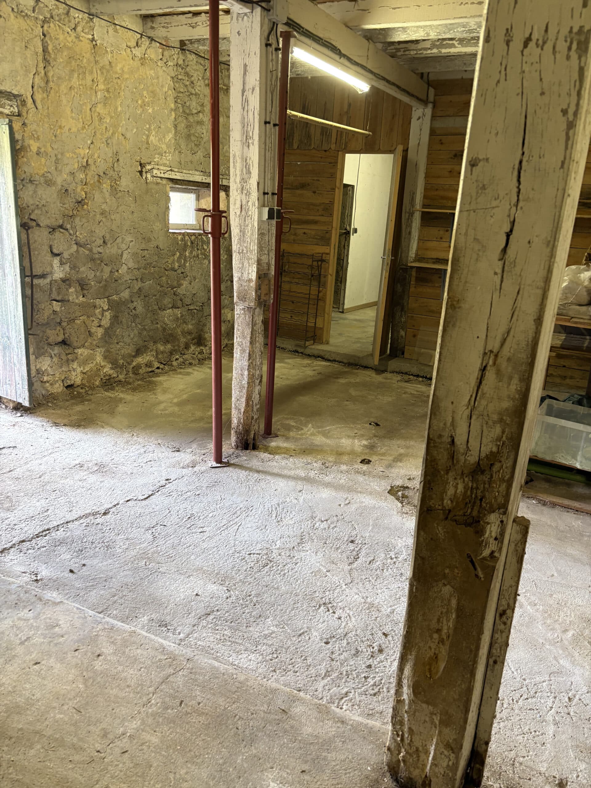 Intérieur rustique, sous-sol ou grange, avec mur en pierre humide, poteau en bois dégradé soutenu par des étais rouges, et sol en béton fissuré.