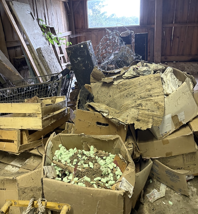 Désordre et Déchets : Cartons Humides dans un Grenier Tas désordonné de cartons humides, caisses en bois et débris, dont des copeaux d'emballage verts, dans un grenier rustique près d'une fenêtre.