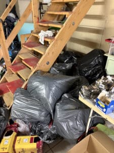 Escalier en bois encombré de sacs poubelles noirs, cartons de vin 'Vieux Papes' et paquets de cigarettes 'Lucky Strike Red'.