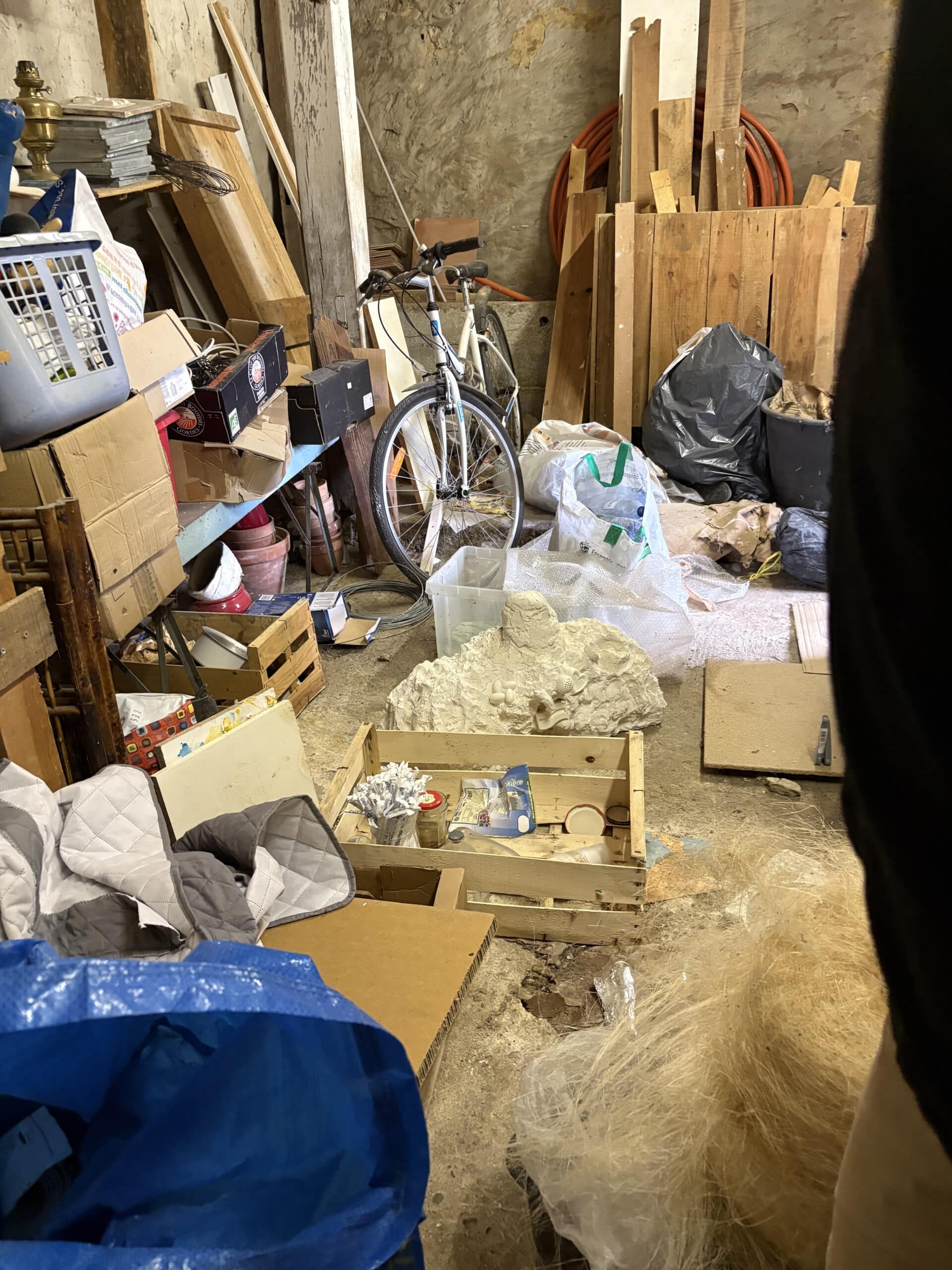 Garage encombré avec un vélo blanc, caisses en bois, sacs poubelles noirs, et gros sac bleu IKEA rempli d'affaires.