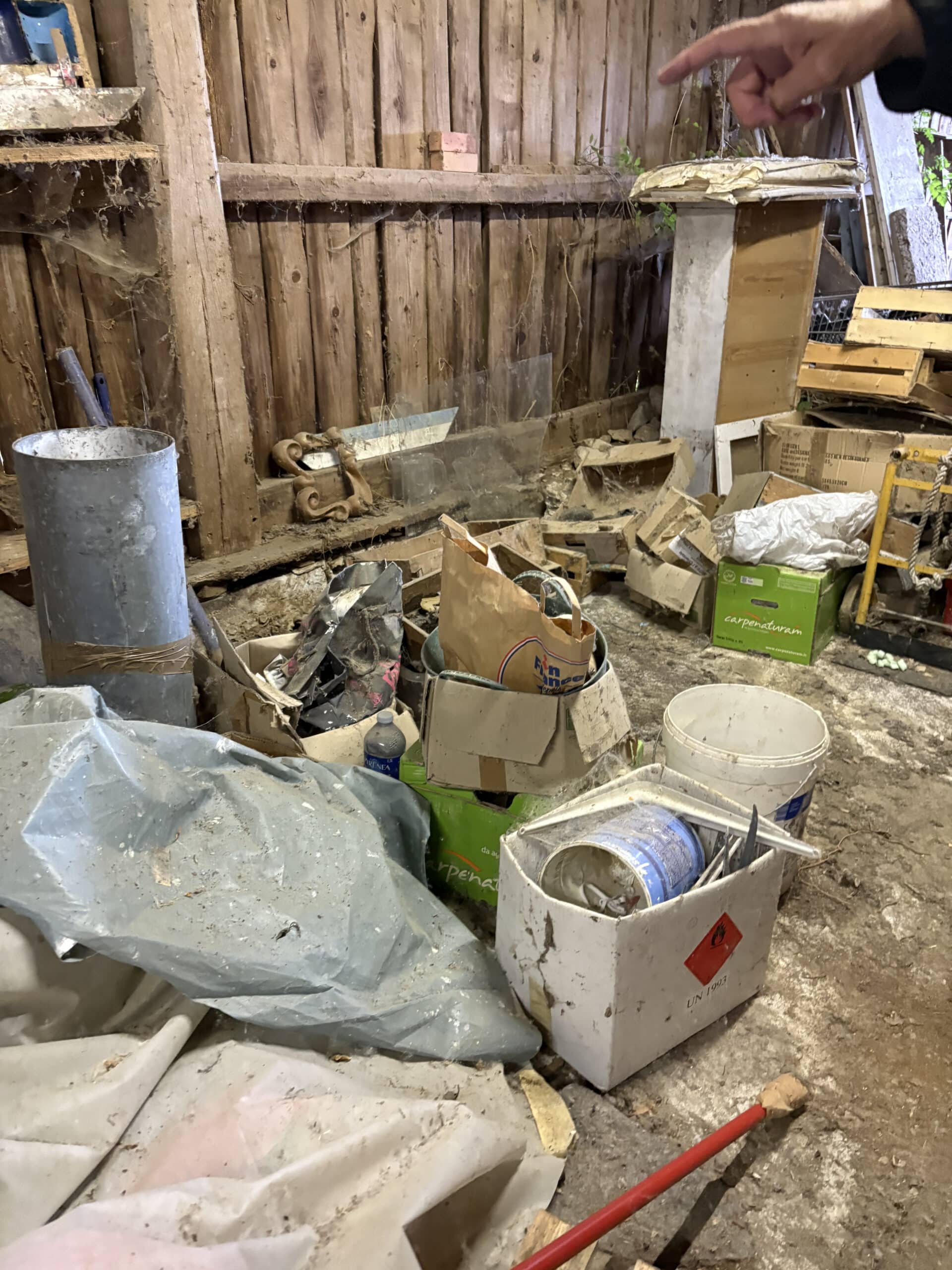 Un homme pointe du doigt dans un hangar encombré de débris, caisses en carton, seaux blancs et une grande bâche grise, fond en bois ancien.
