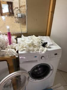 Machine à laver FAURE 7kg, porte ouverte, avec essuie-tout froissé sur le dessus et à côté d'une bouteille rose.