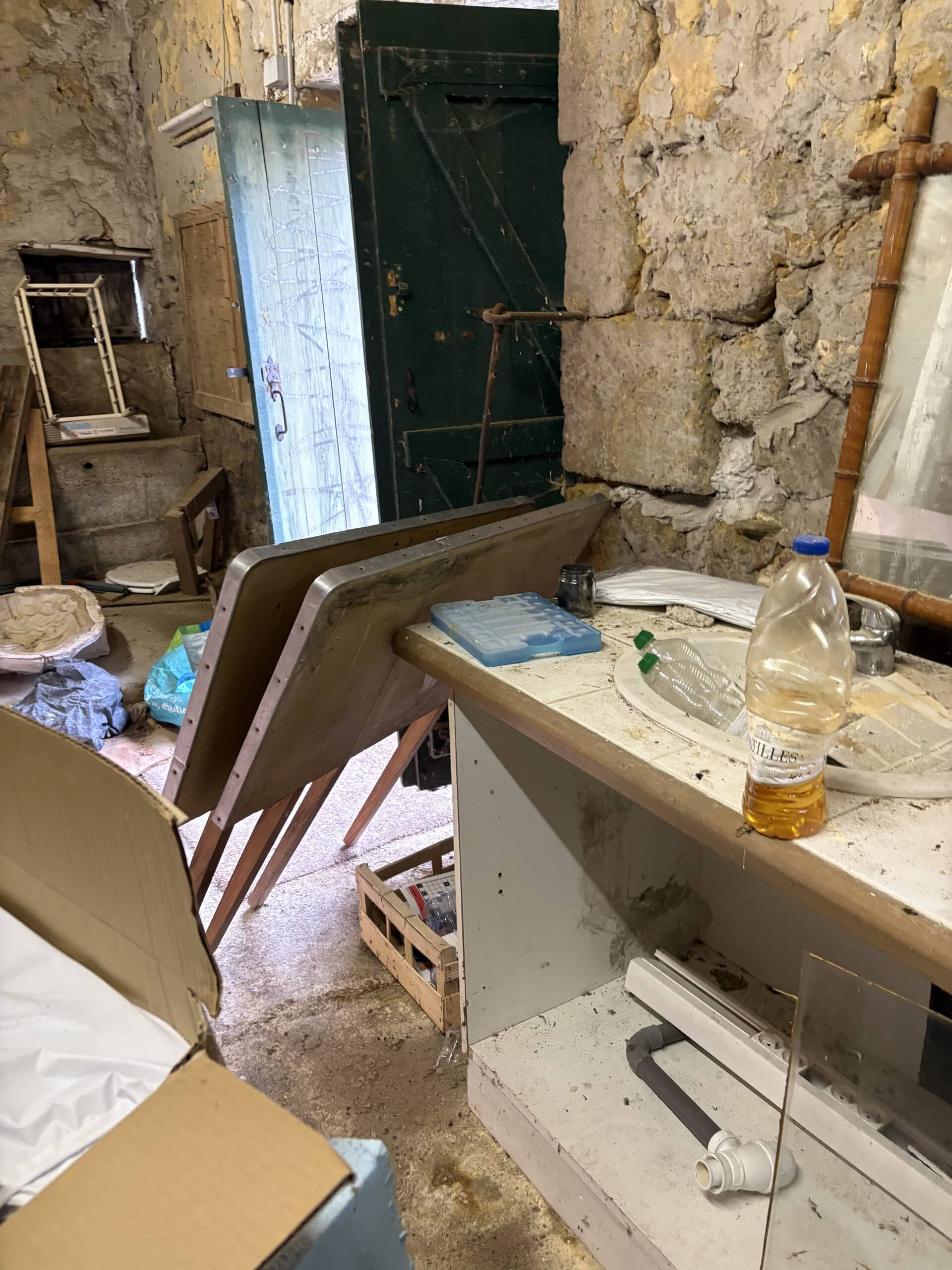 Intérieur de pièce rustique et encombrée avec murs de pierre brute, portes entrouvertes, tables renversées et déchets sur le plan de travail, bouteille "VILLES".
