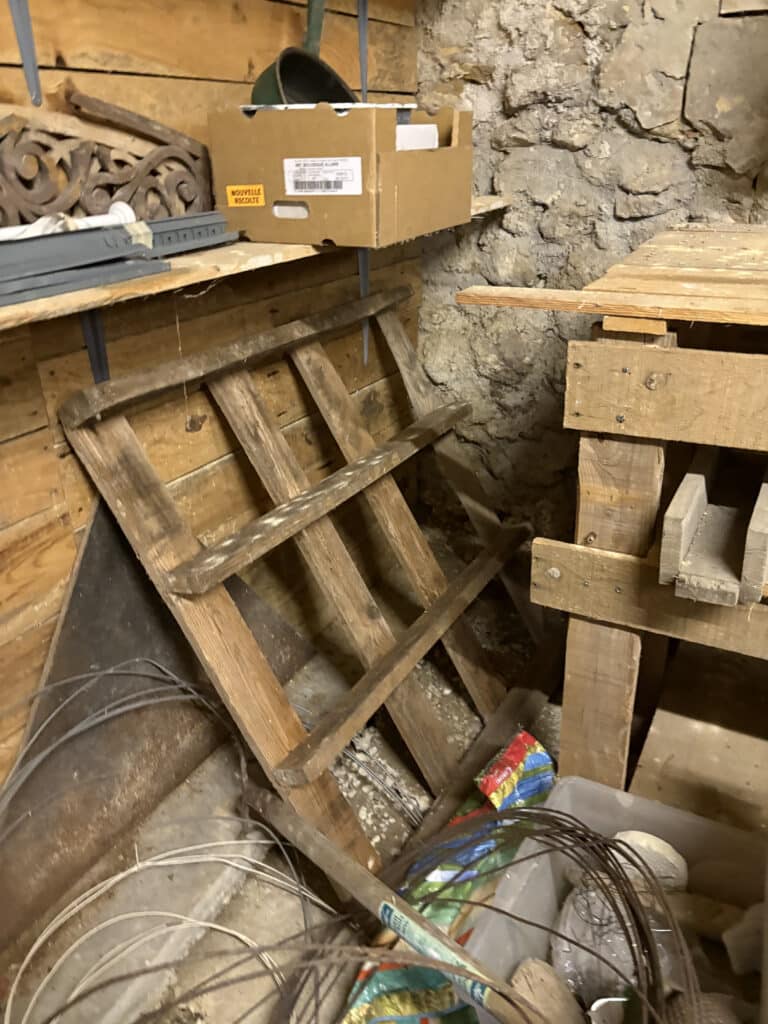 Rangement sous plancher : pierres, bois et outils Rangement sous un plancher avec mur de pierres brutes, vieille échelle en bois inclinée, boîte carton "NOUVELLE RECOLTE", et fils métalliques en vrac.