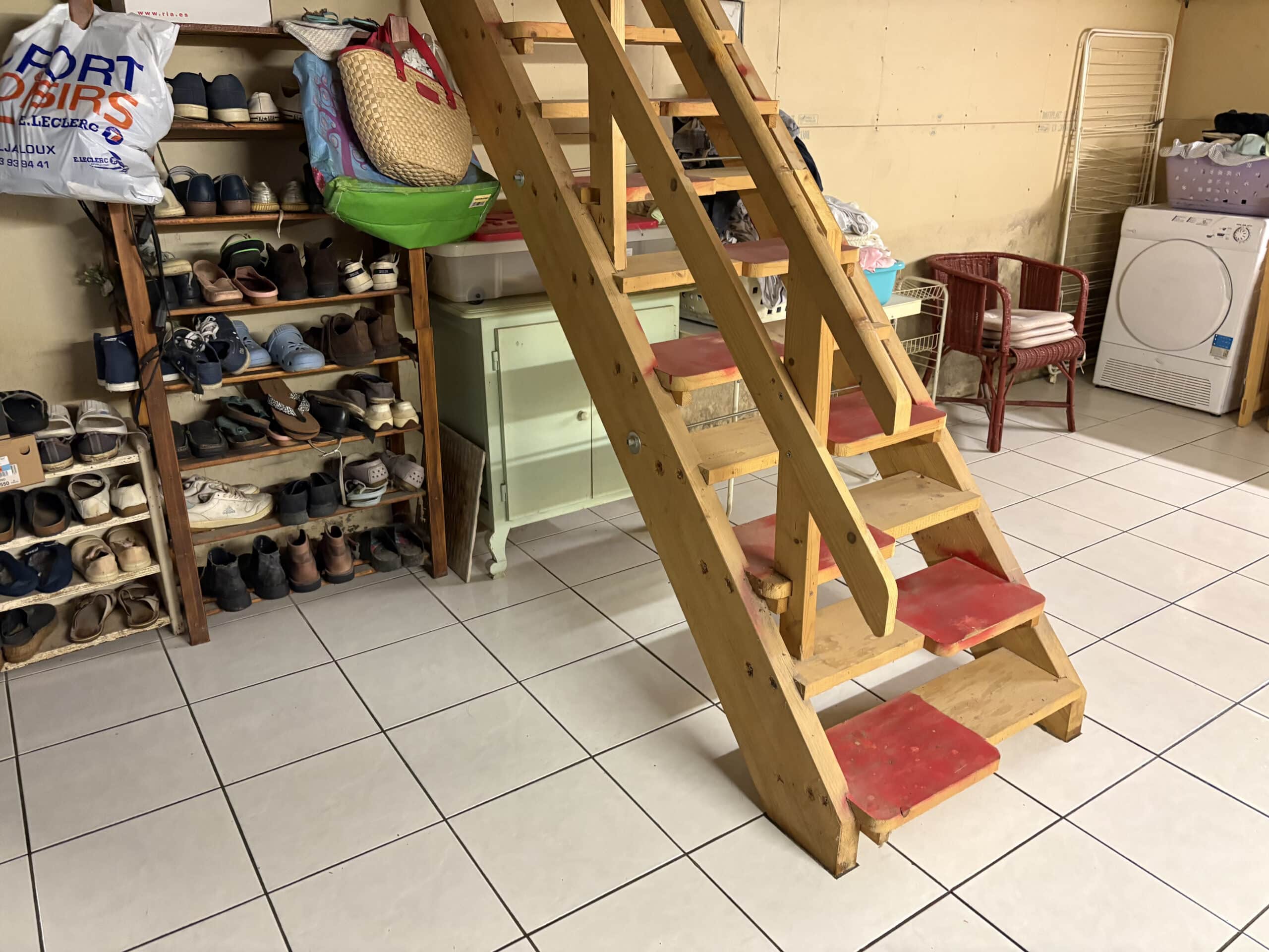 Escalier de bois avec marches rouges, rangements de chaussures et un sèche-linge blanc dans une buanderie carrelée. Sac E.LECLERC visible.