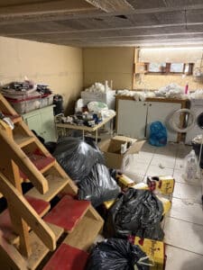 Sous-sol encombré avec sacs poubelles noirs, cartons de 'Vieux Vignes', et cigarettes 'Fumer tue' près d'un escalier et d'appareils ménagers.