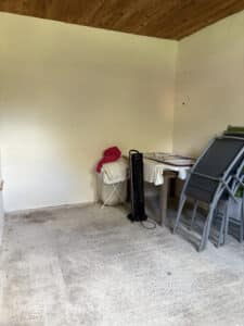 Intérieur brut avec plafond en bois, murs clairs, sol en béton et objets stockés : ventilateur Rowenta, linge et chaises longues pliées.