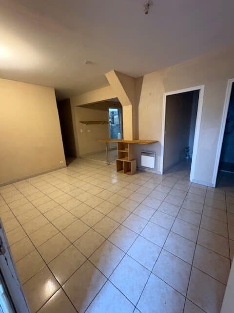 Intérieur vide, pièce principale au carrelage beige brillant. Murs clairs, petite étagère en bois et coin bar. Vue sur une ouverture et deux portes.