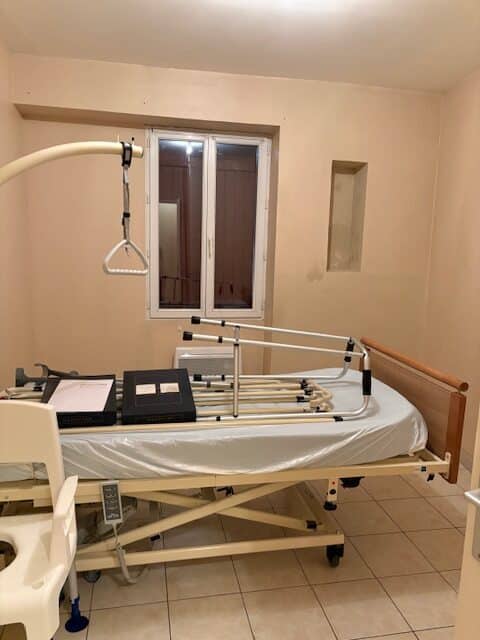 Lit médicalisé réglable avec potence et barrières pliées, béquilles, et chaise de douche dans une chambre de maintien à domicile.