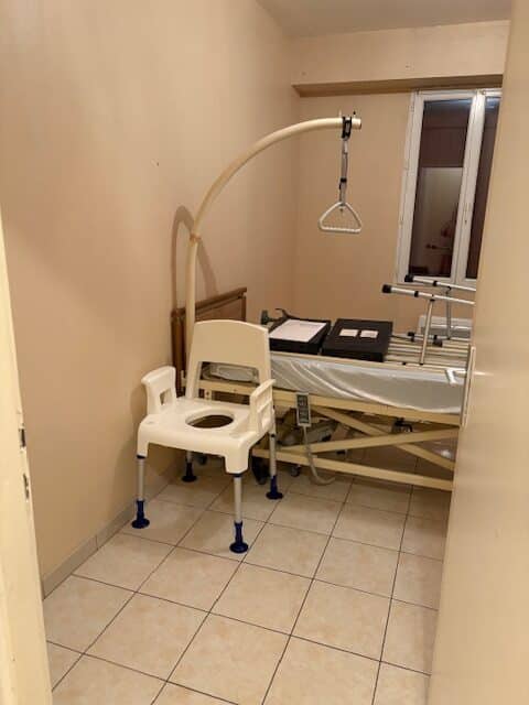 Lit médicalisé et potence dans une chambre claire, avec une chaise de douche/toilettes percée en premier plan. Aménagement PMR.