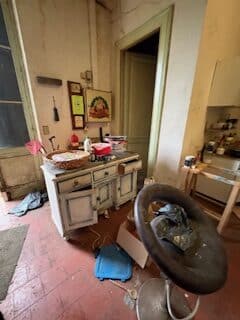 Désordre fascinant : Pièce oubliée et bureau vintage. Intérieur très encombré. Bureau vintage blanchi, chaise ronde pleine de détritus, sol rouge. Thangka bouddhiste au mur d'un atelier négligé.